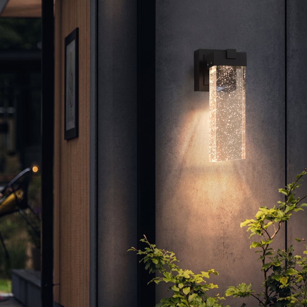 A modern outdoor wall sconce with a rectangular glass bubble column shade, mounted on a dark gray stucco wall and casting a warm light downward, with green plants in the foreground.