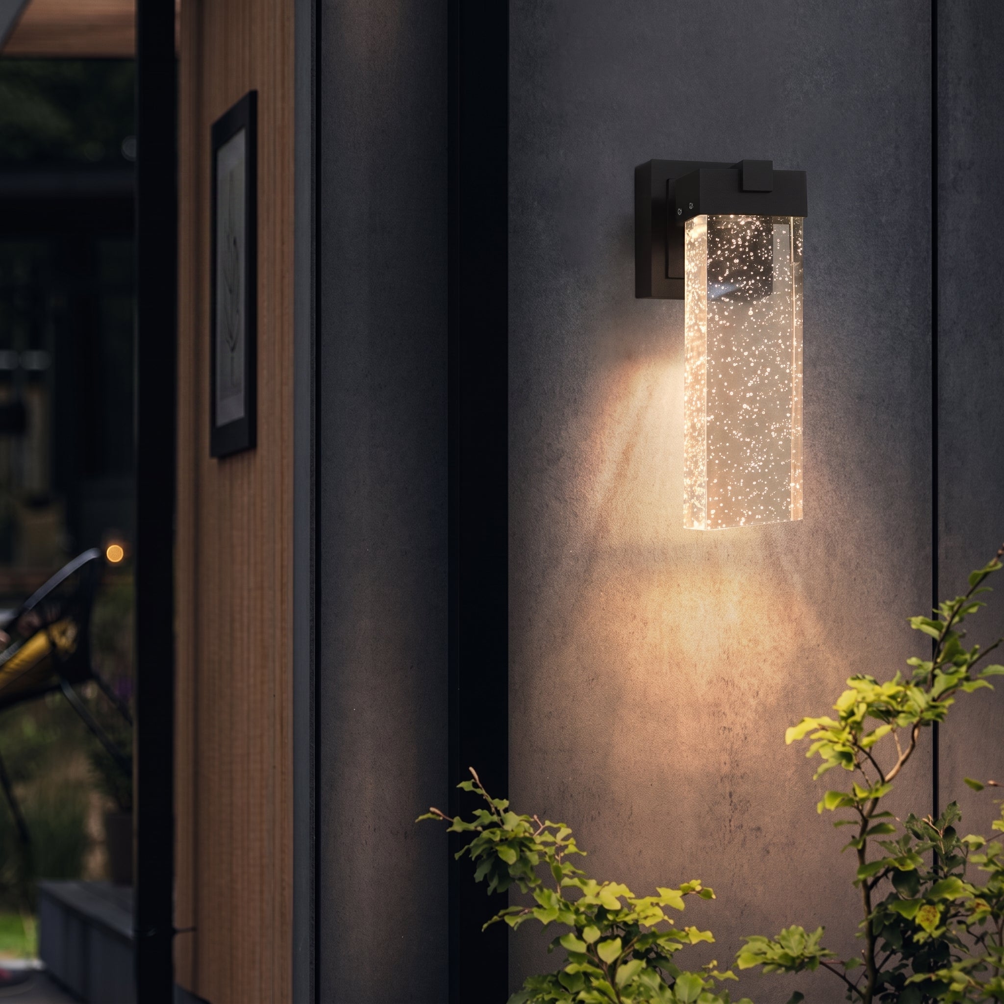A modern outdoor wall sconce with a rectangular glass bubble column shade, mounted on a dark gray stucco wall and casting a warm light downward, with green plants in the foreground.