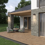 Modern outdoor patio scene with a covered seating area featuring two woven armchairs with green cushions, a small round table, stone columns, a wood slat roof overhang, and wall-mounted light fixtures on a house exterior.
