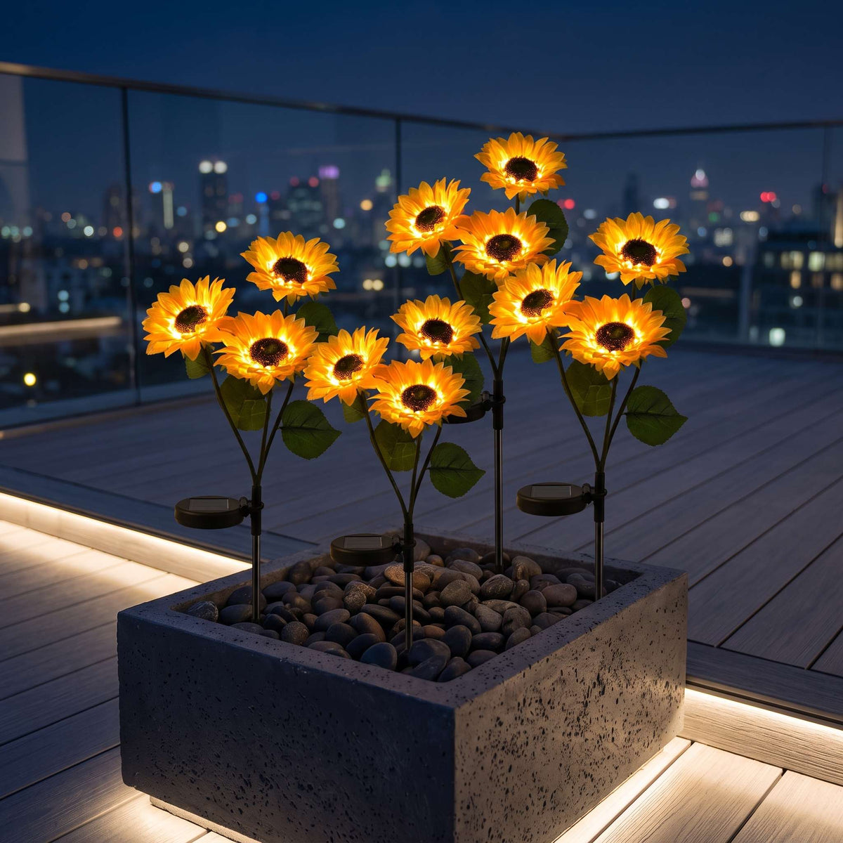 A set of solar-powered LED sunflower lights arranged in a square concrete planter filled with pebbles on a rooftop deck at night, with a blurred city skyline in the background.