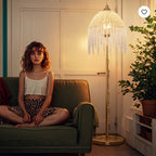 A woman sits cross-legged on an olive green sofa in a warmly lit room, wearing a white camisole and leopard-print bottoms. A tall floor lamp with a gold-toned base and a fringed crystal-like shade stands beside her, next to a potted plant.