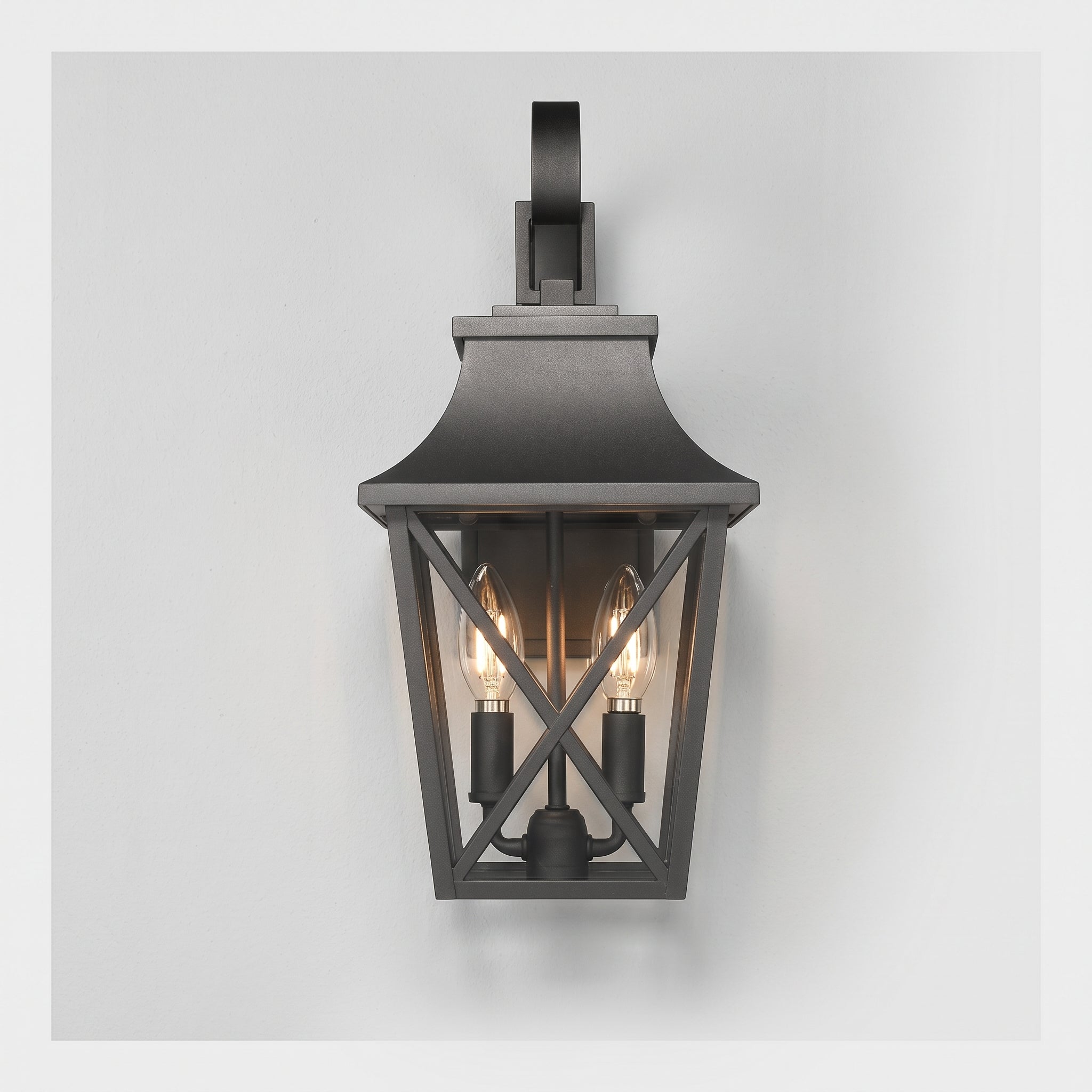 A dark charcoal outdoor wall sconce with a lantern-style frame, two visible warm Edison bulbs, and a curved mounting bracket at the top, shown against a light gray background.