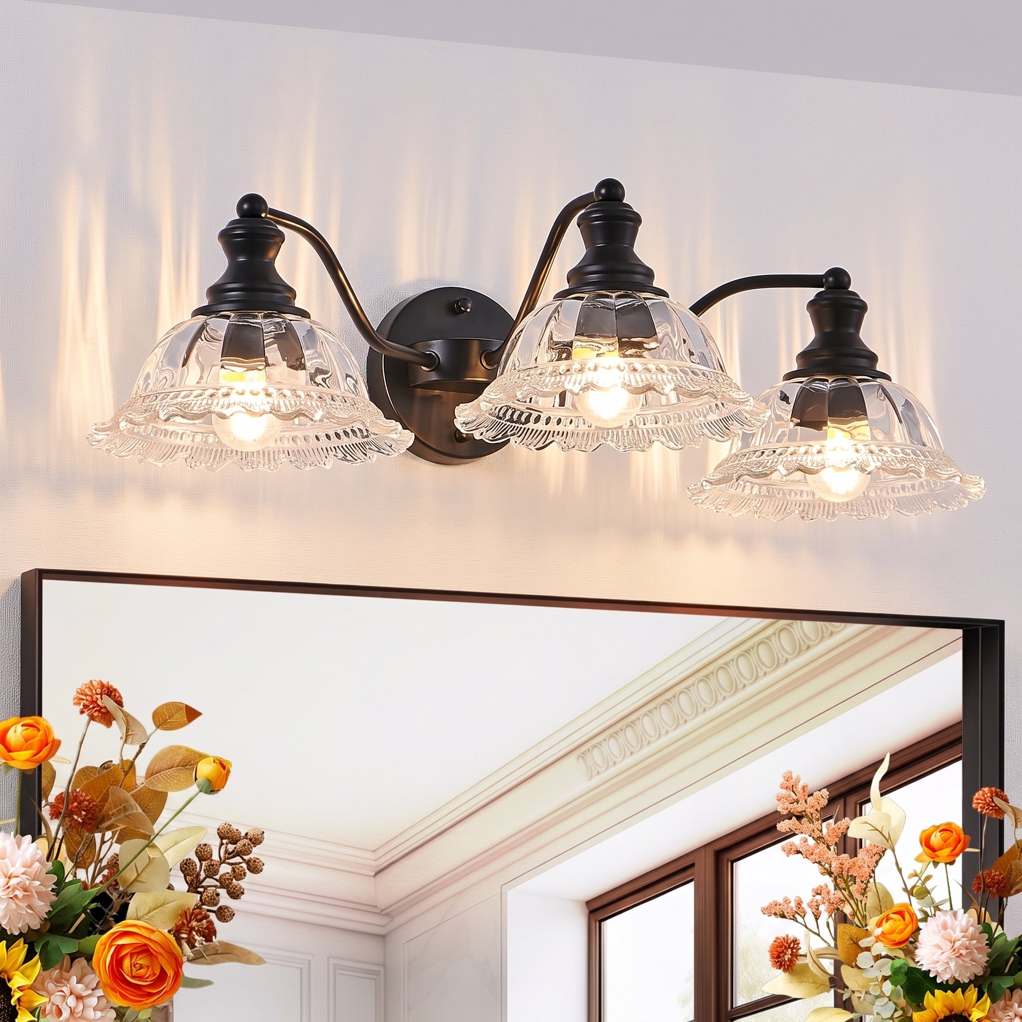 A three-light wall sconce with black metal arms and clear glass shades mounted above a mirror in a bright room. The lights are turned on, casting a warm glow, and floral arrangements are visible in the lower corners of the mirror reflection.