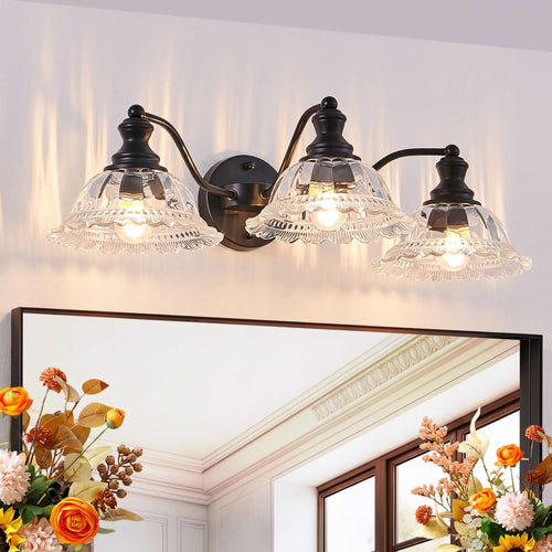 A three-light wall sconce with black metal arms and clear glass shades mounted above a mirror in a bright room. The lights are turned on, casting a warm glow, and floral arrangements are visible in the lower corners of the mirror reflection.