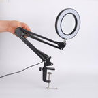 A black adjustable desk lamp with an integrated circular ring light, held by a hand and mounted on a metal clamp with a power cord, shown against a plain white background.