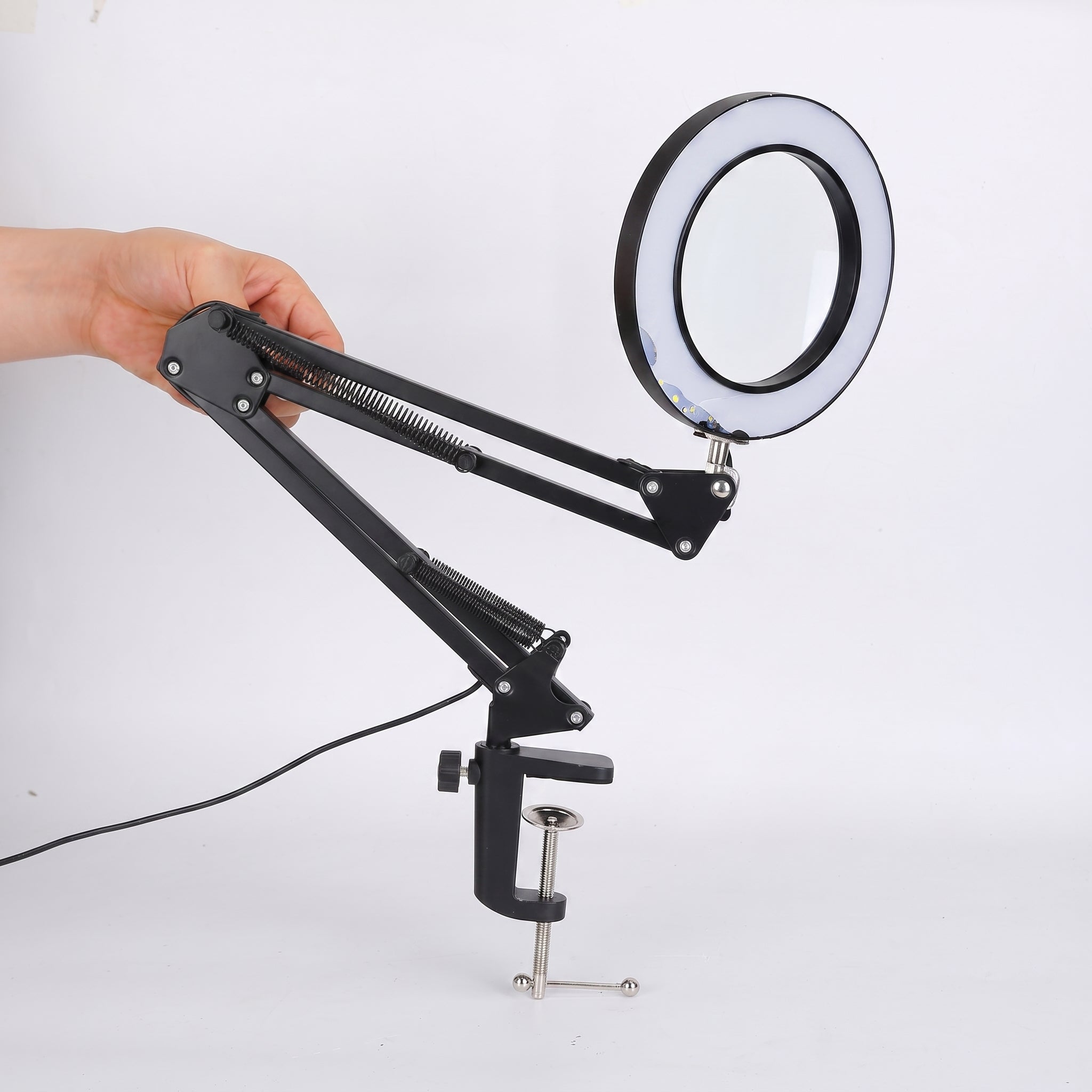A black adjustable desk lamp with an integrated circular ring light, held by a hand and mounted on a metal clamp with a power cord, shown against a plain white background.