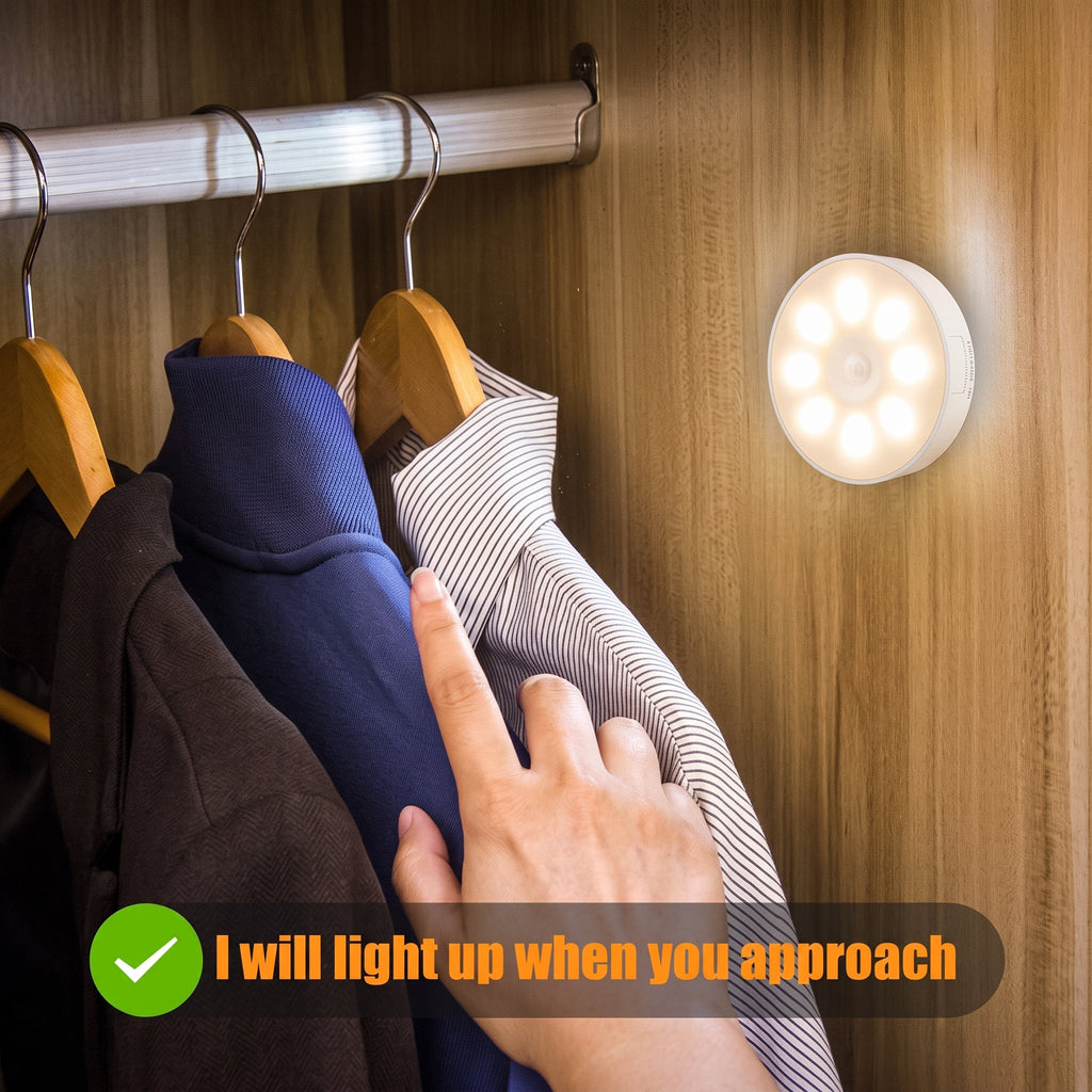 A circular LED closet light glowing on a wooden wardrobe door, with hanging clothes and a hand approaching it to demonstrate the light-up feature. Text overlay reads 'I will light up when you approach'.