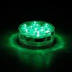 A small circular waterproof LED light glowing green on a black background, with multiple illuminated lenses on top and faint green light rings around it.