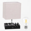 A modern table lamp with a square off-white linen shade, a black power bank-style base with built-in outlets, and a separate clear LED filament bulb shown beside it on a white background.