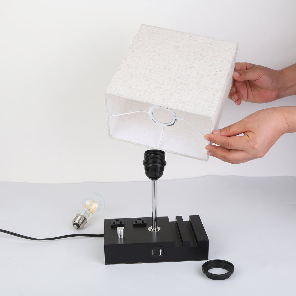 A person's hands are shown holding a white linen-textured table lamp shade above a black multi-port lamp base, demonstrating how to swap the lamp. The setup includes a clear LED filament bulb, a black ring, and a power cord on a clean white background.