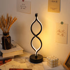 A modern black LED spiral twist table lamp glowing with warm light, placed on a marble desk in a styled home office setting with books, a candle, and wall art.