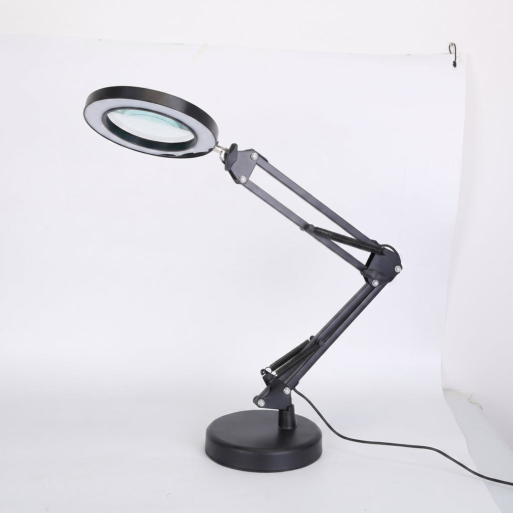A black adjustable desk lamp with an integrated magnifying glass on the lamp head, mounted on a circular base with an articulated arm, shown against a white studio background.