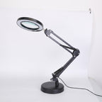 A black adjustable desk lamp with an integrated magnifying glass on the lamp head, mounted on a circular base with an articulated arm, shown against a white studio background.