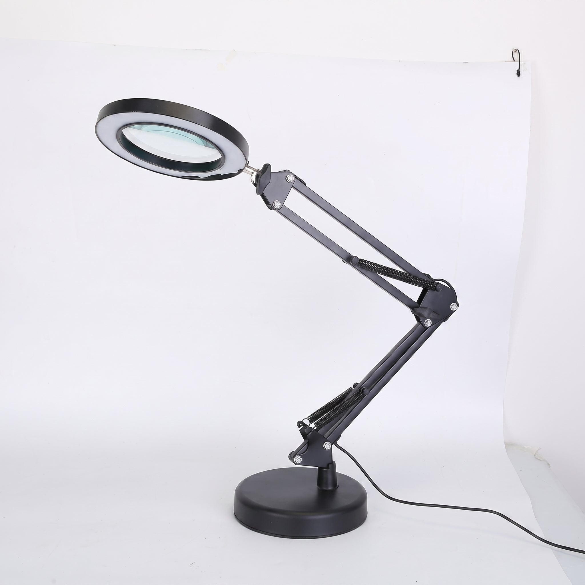 A black adjustable desk lamp with an integrated magnifying glass on the lamp head, mounted on a circular base with an articulated arm, shown against a white studio background.