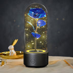 A glass dome keepsake flower lamp featuring a blue rose with gold fairy lights inside, set on a black base with a power button, displayed on a table with a dark gray background.