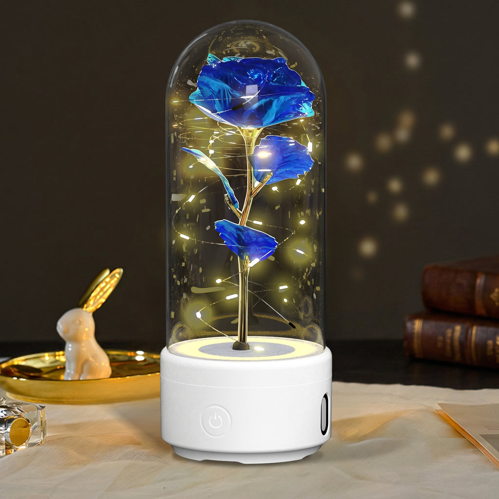 A preserved blue rose under a clear glass dome with warm fairy lights inside, displayed on a white base on a tabletop with soft-focus decorative props in the background.