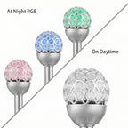 Three solar garden post lights with crystal-embellished spherical heads are shown on a white background, split diagonally by a gray line. The left side reads 'At Night RGB' and shows the lights colored pink, blue, and green.