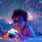 A young child wearing blue swim goggles lies on sand in the ocean, looking at a glowing sea urchin-shaped night light that projects colorful underwater patterns across the water.