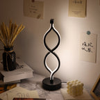 A modern black LED table lamp with a double-swoosh geometric design and a circular base, illuminated with a warm white glow on a marble desk in a styled room setting.