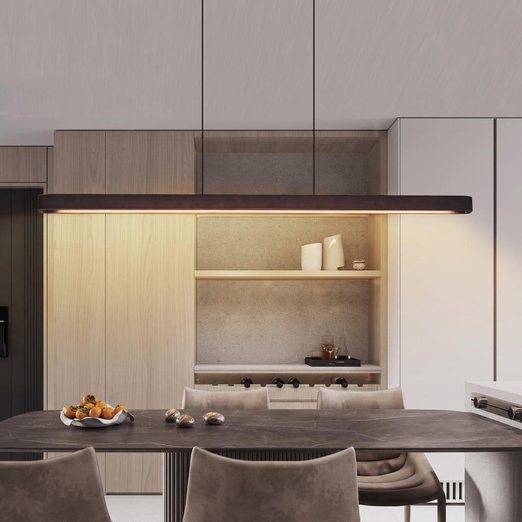 A modern kitchen interior with a dark rectangular pendant light hanging above a dining table and chairs. The space features light wood cabinetry, open shelving with white ceramic vessels, a textured stone backsplash, and a dark wood tabletop.