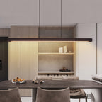 A modern kitchen interior with a dark rectangular pendant light hanging above a dining table and chairs. The space features light wood cabinetry, open shelving with white ceramic vessels, a textured stone backsplash, and a dark wood tabletop.