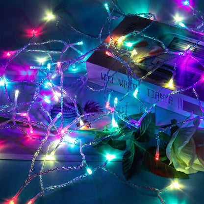 Multicolor LED fairy string lights draped over a stack of books and green leaves, creating a colorful, festive atmosphere.