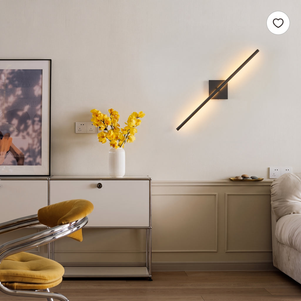 A modern wall-mounted linear light fixture with a black metal frame and warm glowing bulb, mounted on a beige wall in a styled living room setting.