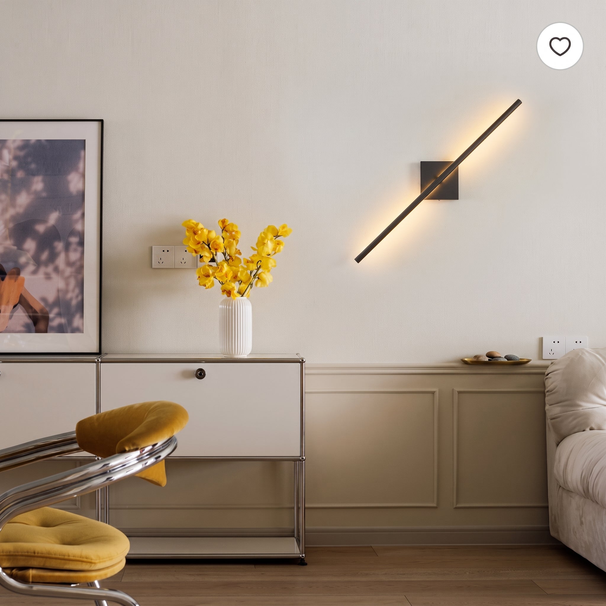A modern wall-mounted linear light fixture with a black metal frame and warm glowing bulb, mounted on a beige wall in a styled living room setting.