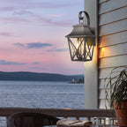 A classic outdoor wall lantern light fixture mounted on a house exterior, casting a warm glow over a waterfront patio scene at dusk with a lake, distant hills, and a potted plant visible.