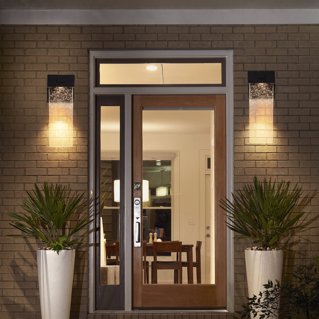 A modern double front door with glass panels and a transom window, set in a brick entryway at night. Two rectangular wall sconces glow on either side, and potted plants sit on both sides of the door.