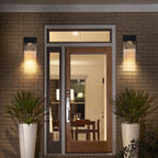A modern double front door with glass panels and a transom window, set in a brick entryway at night. Two rectangular wall sconces glow on either side, and potted plants sit on both sides of the door.