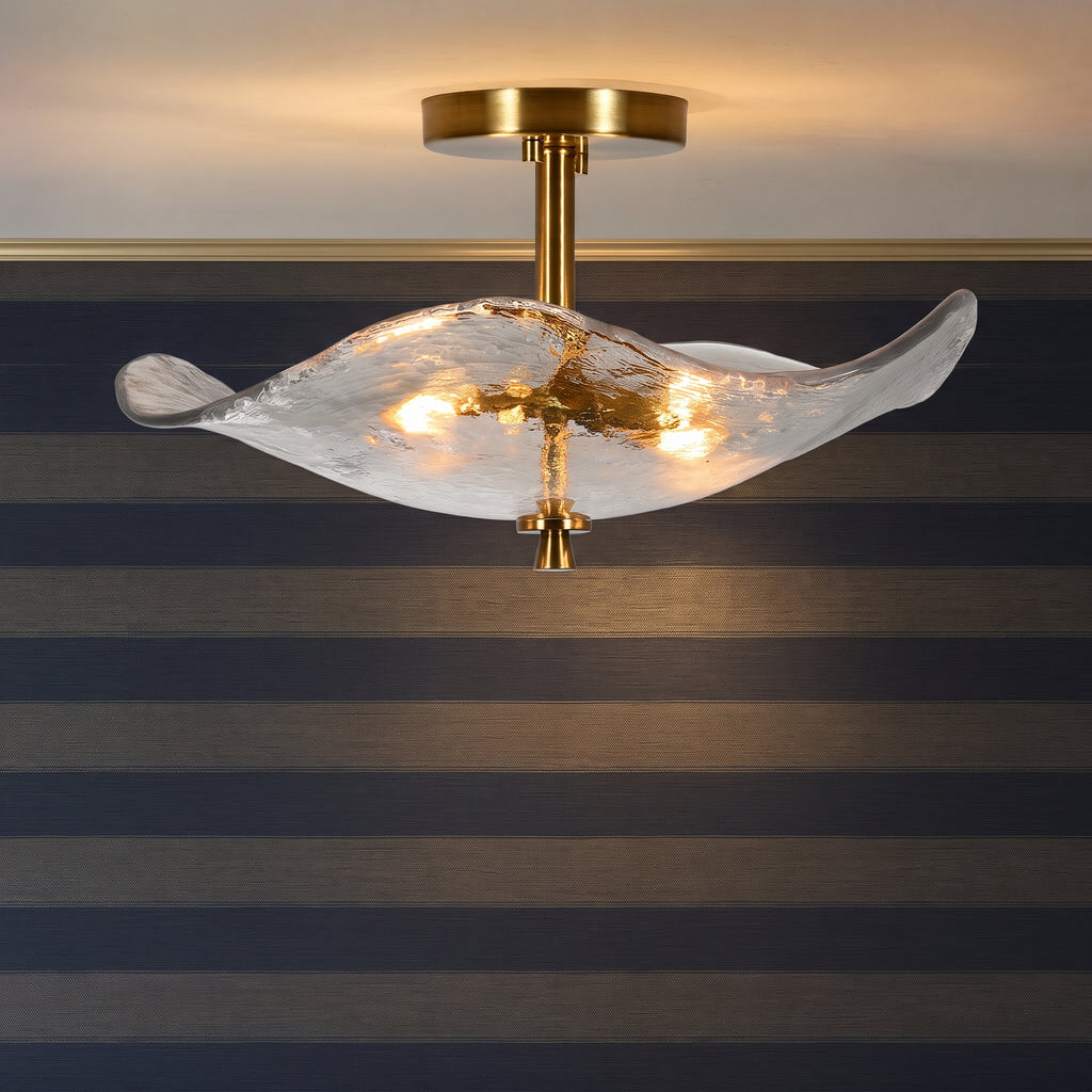 A modern semi-flush mount ceiling light fixture with a wavy, translucent textured glass shade and a brushed gold brass ceiling canopy and central stem, shown illuminated against a striped wall.
