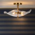 A modern semi-flush mount ceiling light fixture with a wavy, translucent textured glass shade and a brushed gold brass ceiling canopy and central stem, shown illuminated against a striped wall.