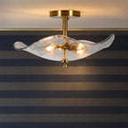A modern semi-flush mount ceiling light fixture with a wavy, translucent textured glass shade and a brushed gold brass ceiling canopy and central stem, shown illuminated against a striped wall.