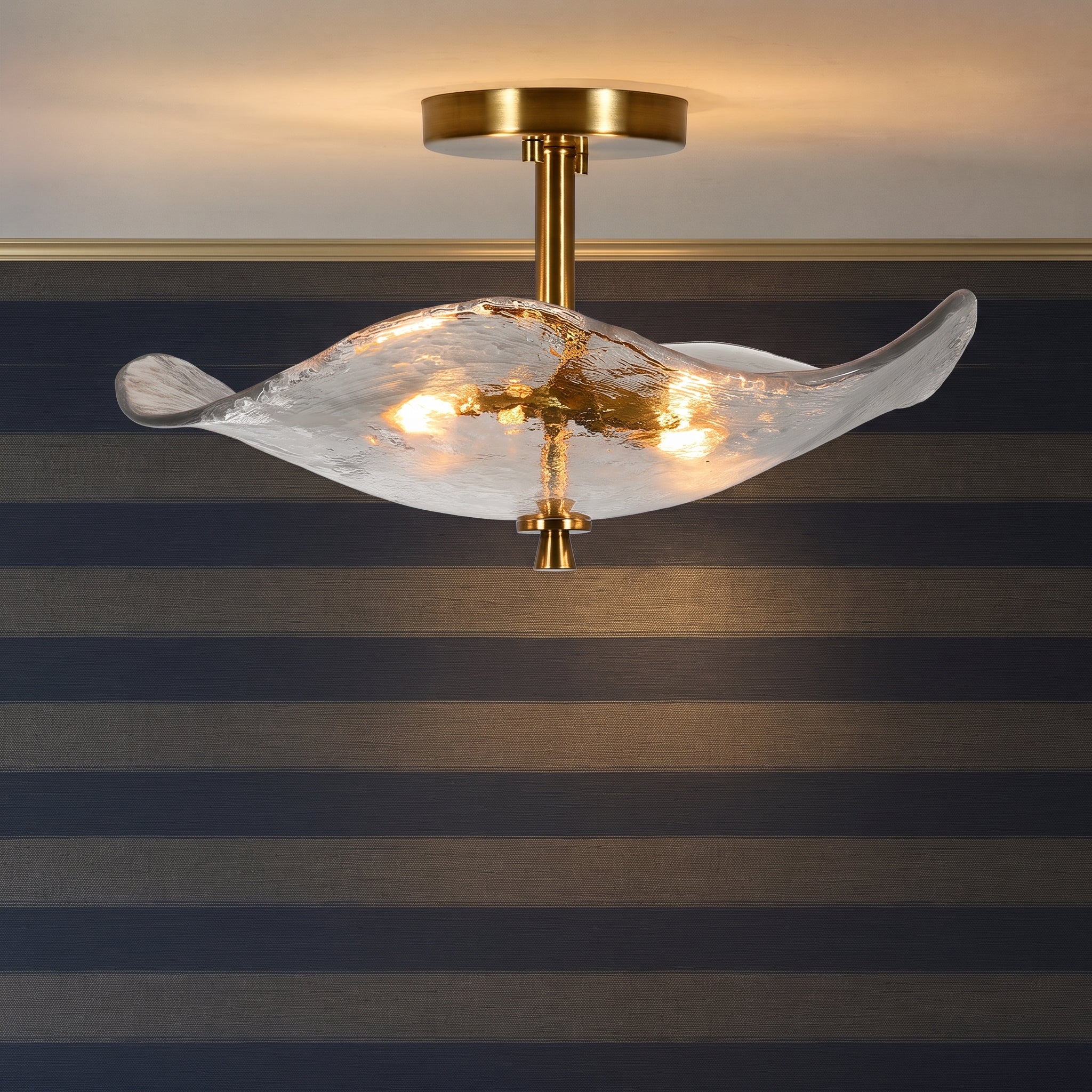 A modern semi-flush mount ceiling light fixture with a wavy, translucent textured glass shade and a brushed gold brass ceiling canopy and central stem, shown illuminated against a striped wall.