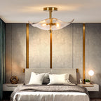 A modern ceiling light fixture with a gold-toned brass frame and a translucent, petal-like glass shade is mounted above a bed in a contemporary bedroom with textured gray walls and gold vertical accents.