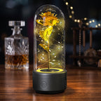A preserved yellow flower inside a clear glass dome lamp with a dark gray base, lit with warm white spiral fairy lights on a wooden table. A blurred glass decanter and string lights appear in the background.