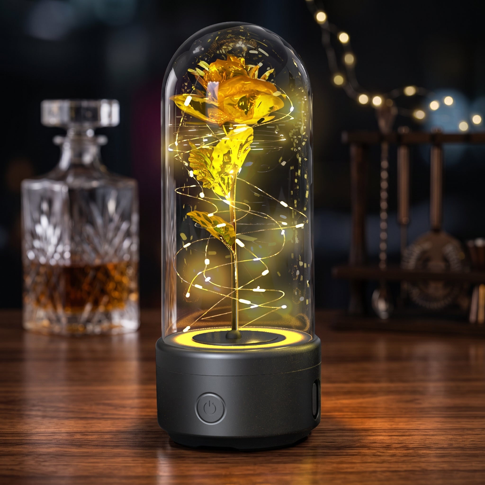 A preserved yellow flower inside a clear glass dome lamp with a dark gray base, lit with warm white spiral fairy lights on a wooden table. A blurred glass decanter and string lights appear in the background.