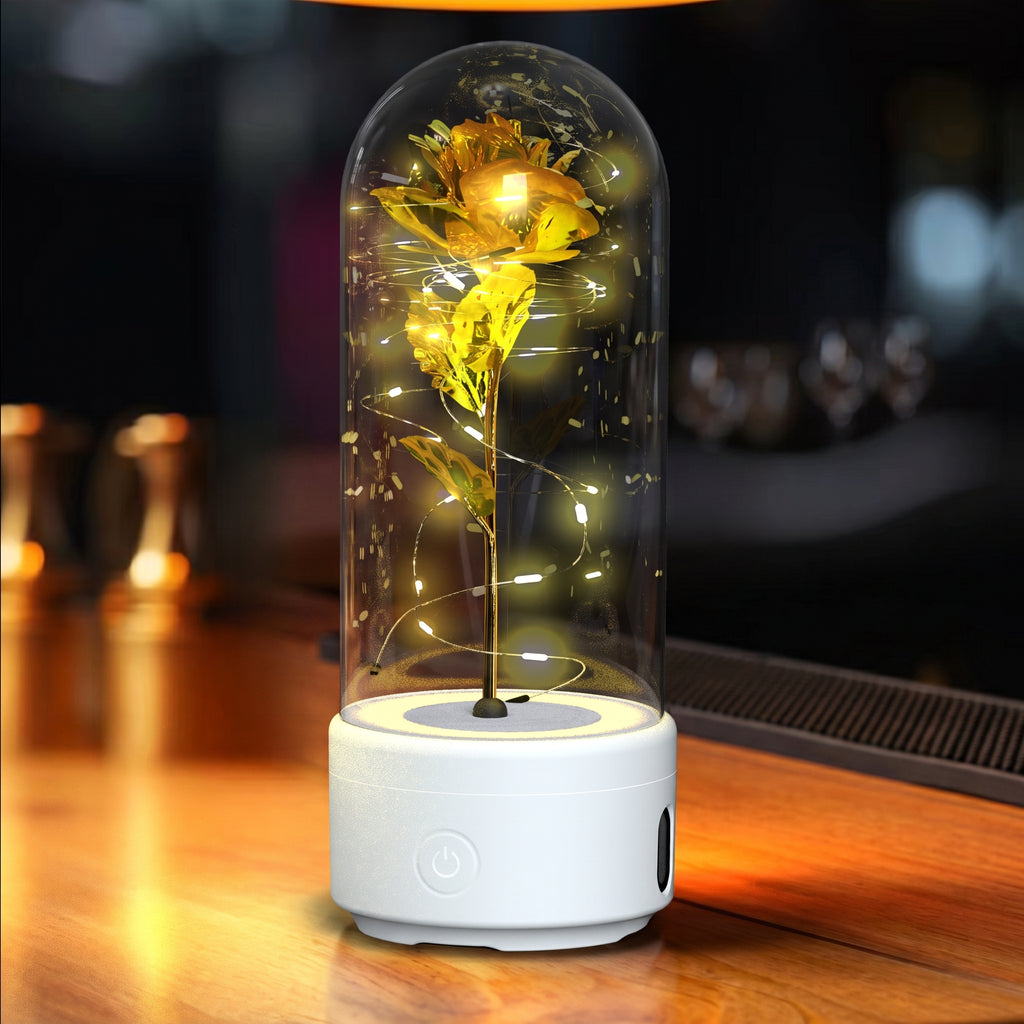 A preserved yellow rose inside a clear glass dome on a white cylindrical base, lit with warm fairy lights, set on a wooden surface in a dimly lit environment.