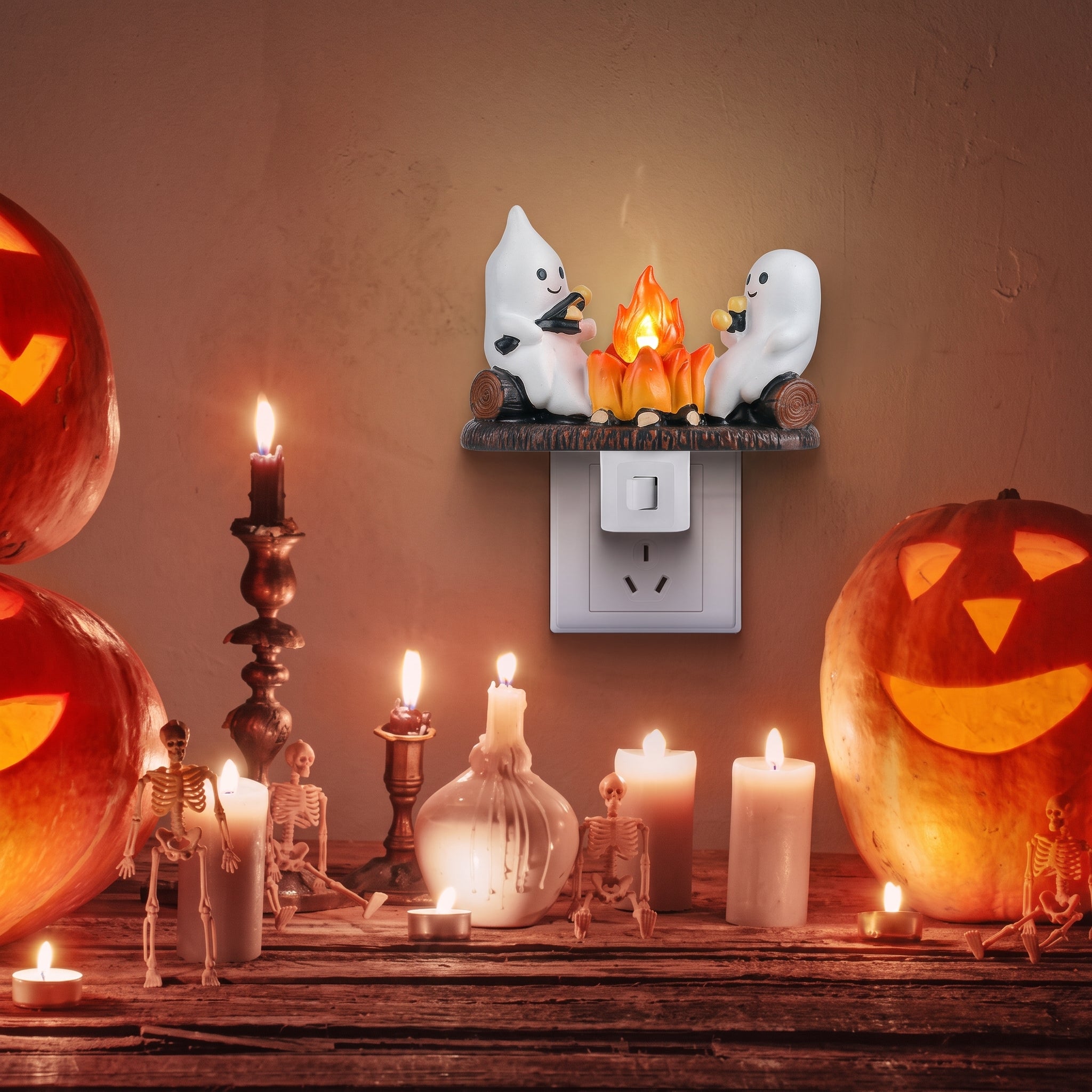 A Halloween-themed night light plug-in featuring two white ghosts sitting around a small orange flame, mounted on a wall above a white outlet. The scene is styled with carved pumpkins, candles, and skeleton props on a wooden surface below.