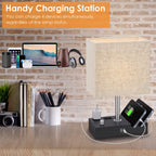 A desk charging station setup featuring a table lamp with a beige fabric shade and a black base that includes built-in chargers and cables, shown on a wooden desk against a brick wall. An orange text banner at the top reads 'Handy Charging Station' with additional descriptive copy about charging four devices simultaneously.