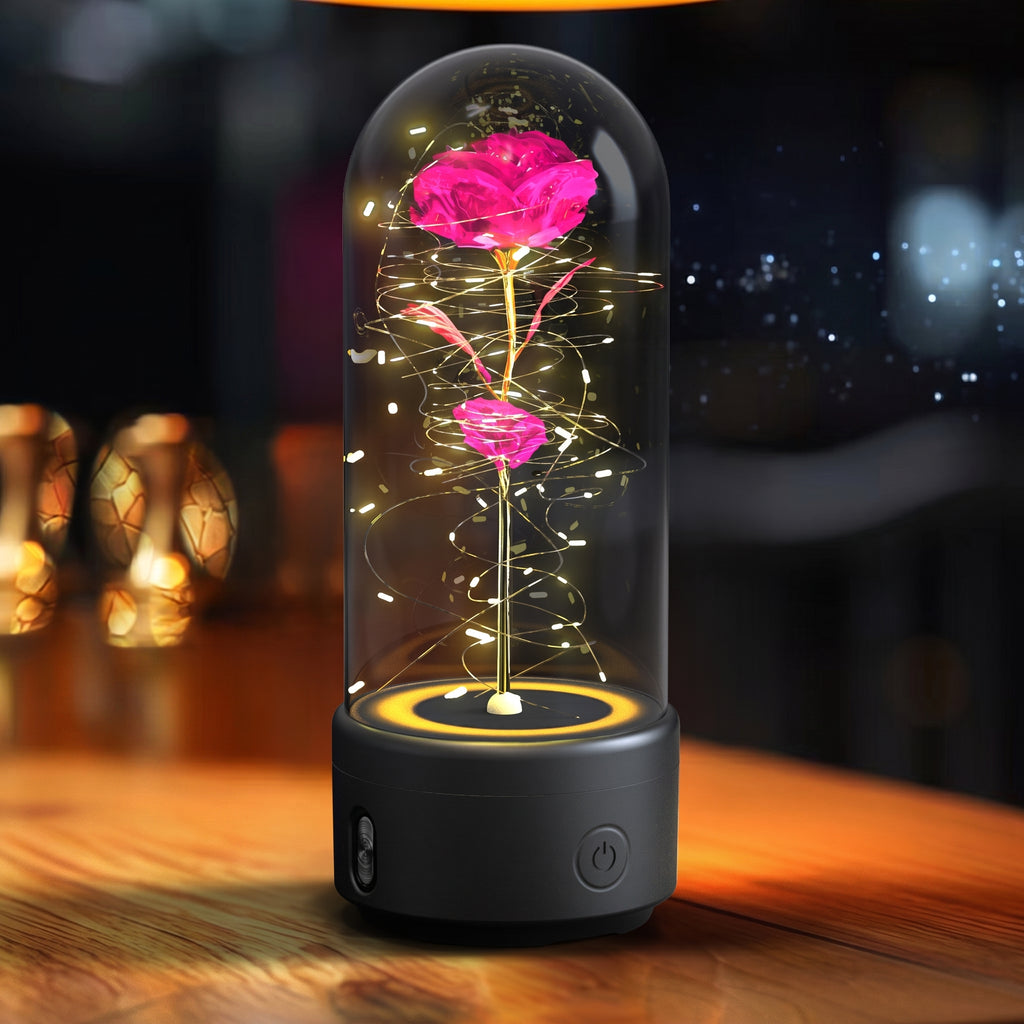 A preserved pink rose inside a clear glass dome lamp with warm white LED fairy lights and a black base, shown on a wooden table in a softly blurred indoor setting.