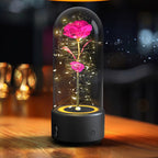 A preserved pink rose inside a clear glass dome lamp with warm white LED fairy lights and a black base, shown on a wooden table in a softly blurred indoor setting.