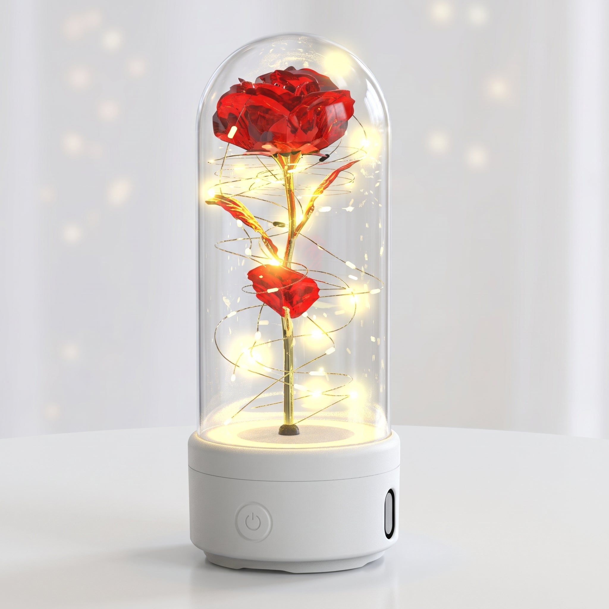 A preserved red rose in a clear glass dome with warm white fairy lights wrapped around the stem, sitting on a white cylindrical base against a softly lit white background.