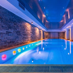 An indoor swimming pool with bright blue water, a stone wall on one side, and floating colorful light spheres along the edge. The space is illuminated by warm wall lighting and recessed lights in a blue ceiling.