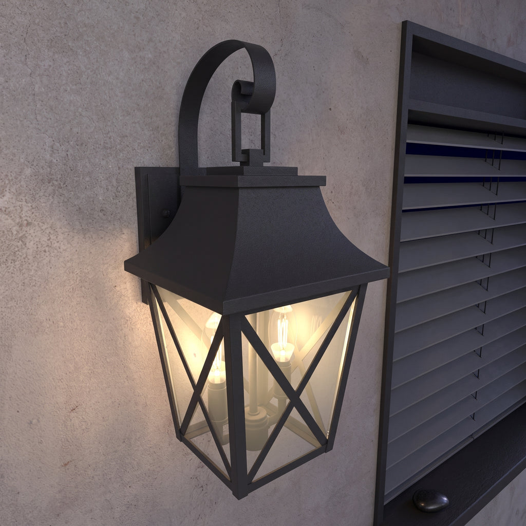 A black outdoor wall lantern light fixture mounted on a textured concrete wall, shown illuminated with a warm glow. The lantern has a geometric frame and is positioned next to a dark gray window or shutter.