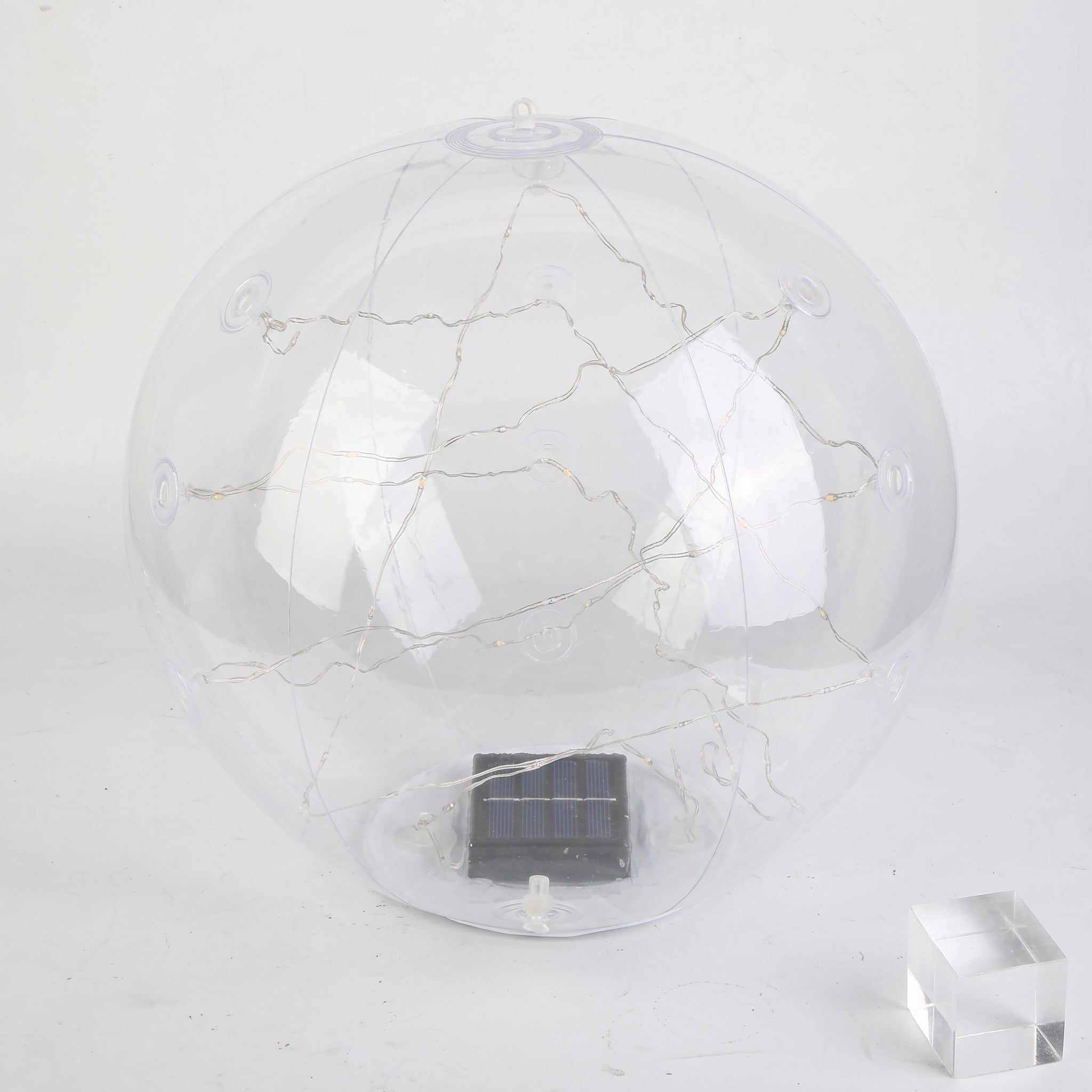 A clear spherical solar globe lamp with internal warm white string lights and a small dark solar panel at the base, shown on a plain light background with a small transparent block prop.