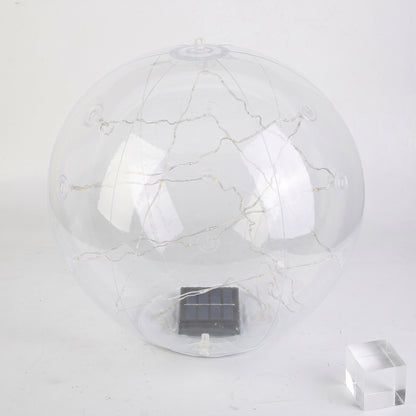 A clear spherical solar globe lamp with internal warm white string lights and a small dark solar panel at the base, shown on a plain light background with a small transparent block prop.