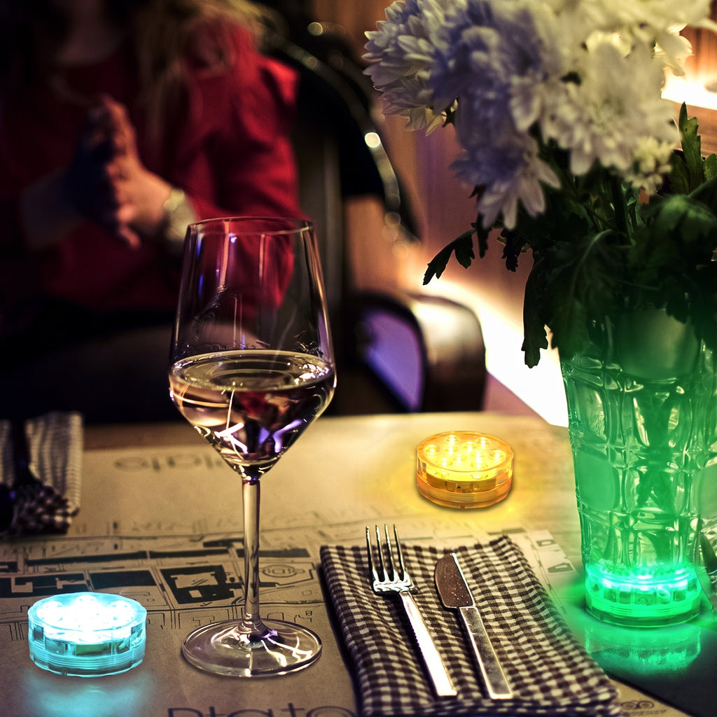 A dimly lit dining table setting with a wine glass partially filled with white wine, a black-and-white checkered napkin with silver fork and knife, a glowing blue candle holder, a yellow candle, and a green illuminated vase of flowers.