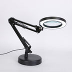 A black adjustable desk magnifier lamp with a circular LED ring light and a large viewing lens, mounted on a round base with a power cord, shown against a plain white background.